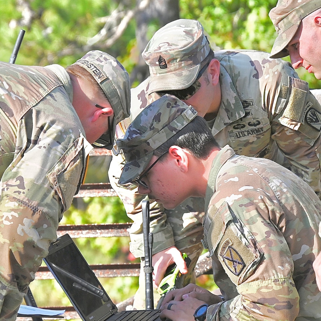 Three Soldiers outdoors squatting around a laptop computer as one Soldier operates it.