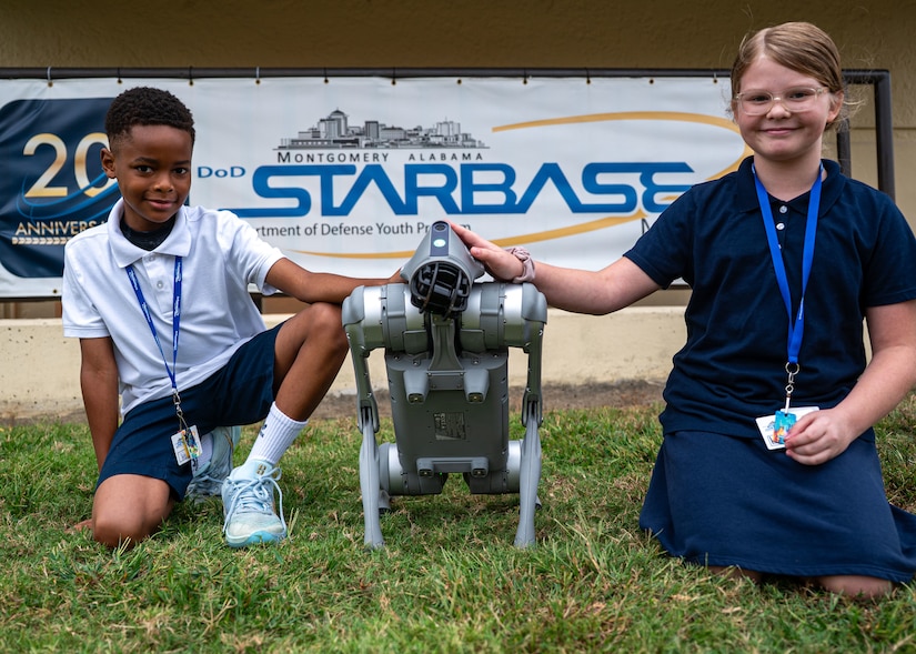 Two children wearing casual attire kneel while posing for a photo with a robotic dog.