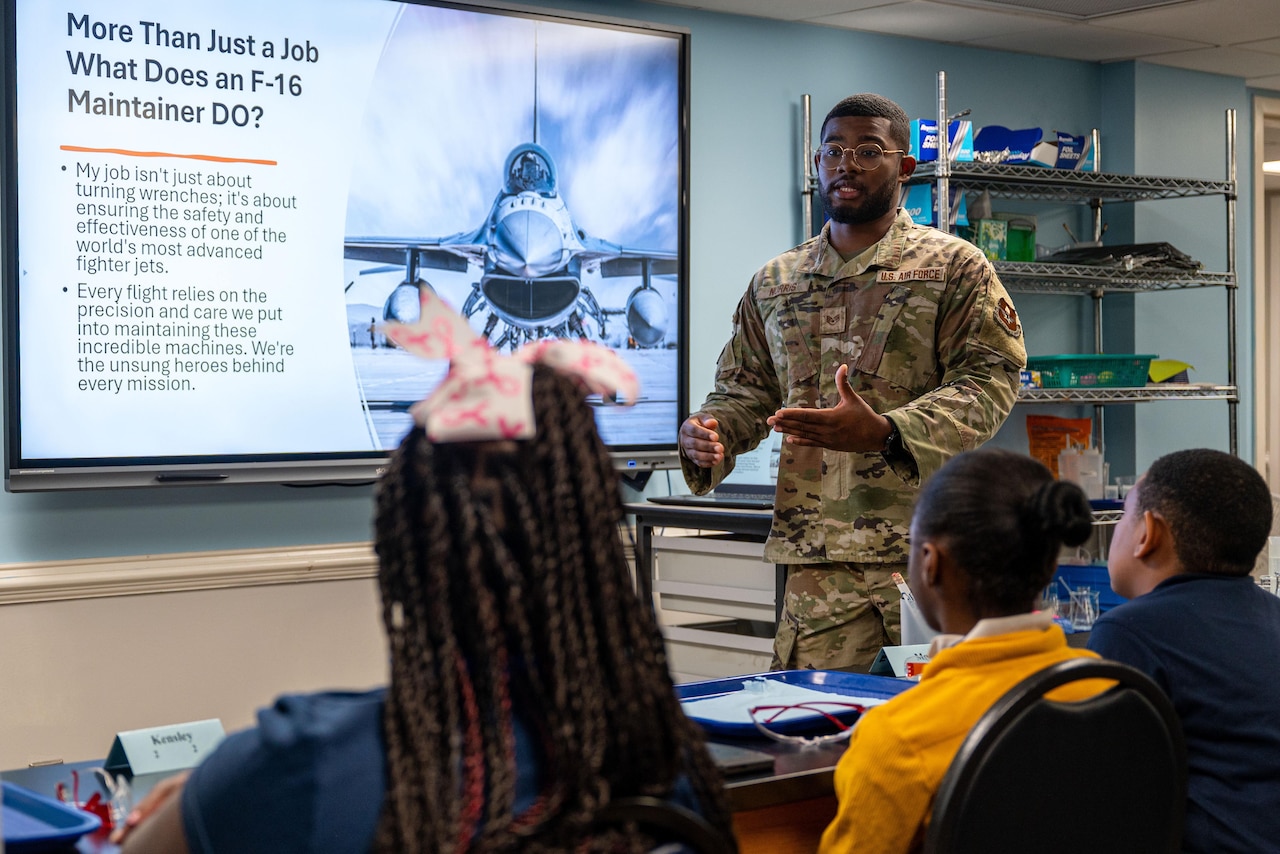 An airman in a camouflage uniform stands at the front of a room while giving instructions to a group of seated schoolchildren. Behind the airman is a monitor with a slide featuring a photo of a military fighter jet.