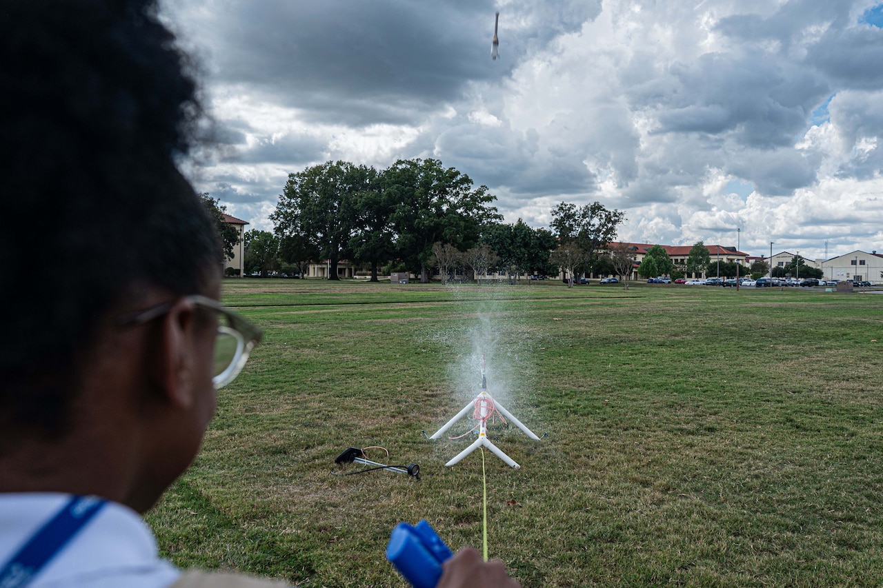 A youth stands in the foreground and remotely operates a model rocket that launches from the ground.
