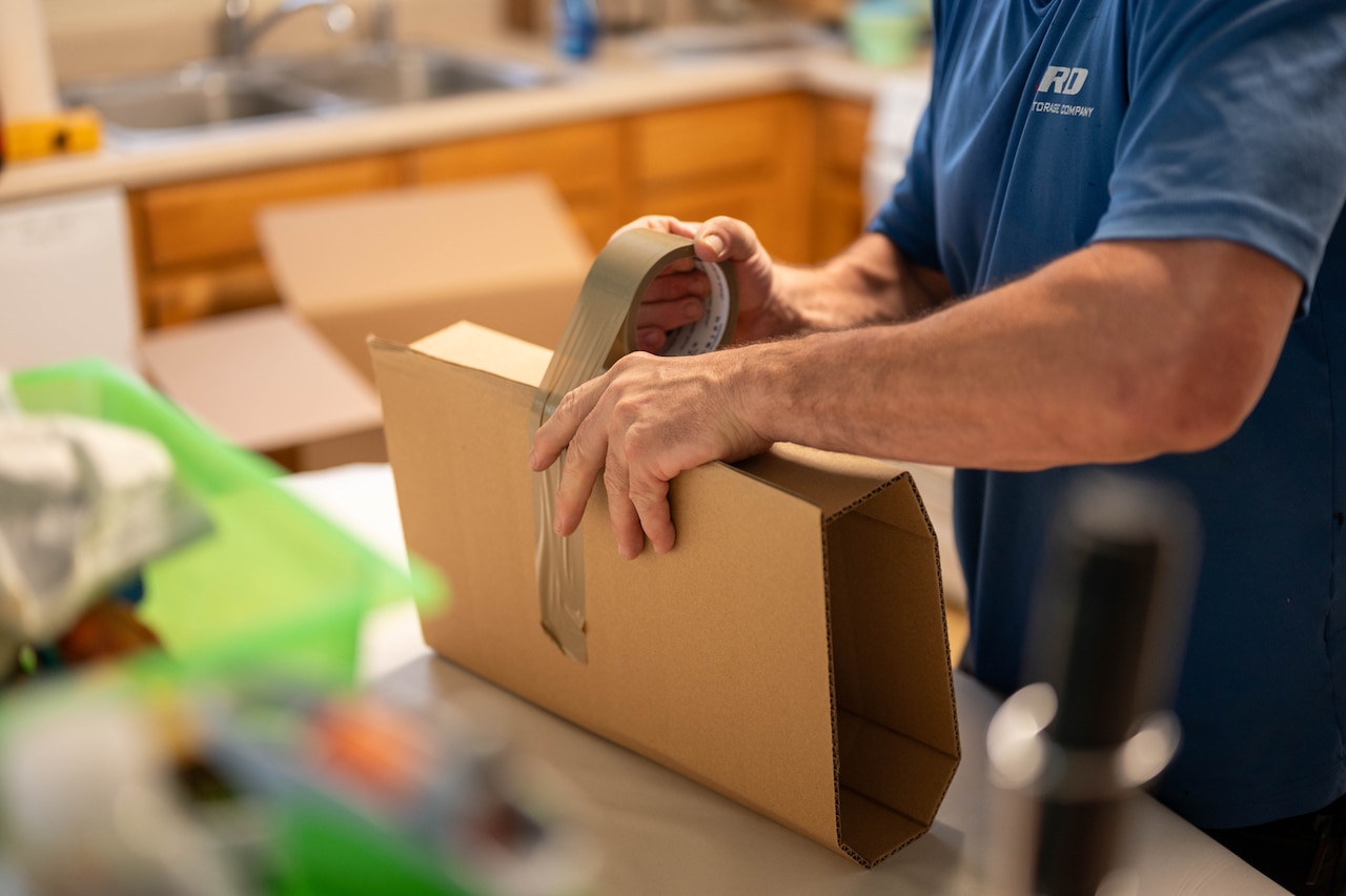 A person in casual attire wraps an object in packing cardboard while standing in a kitchen.