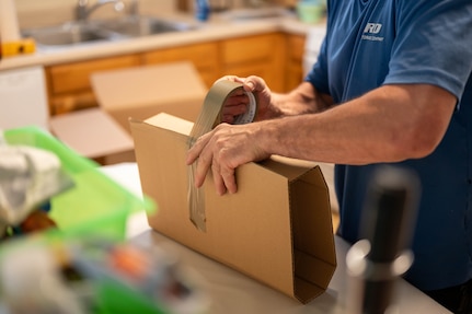 A person in casual attire wraps an object in packing cardboard while standing in a kitchen.