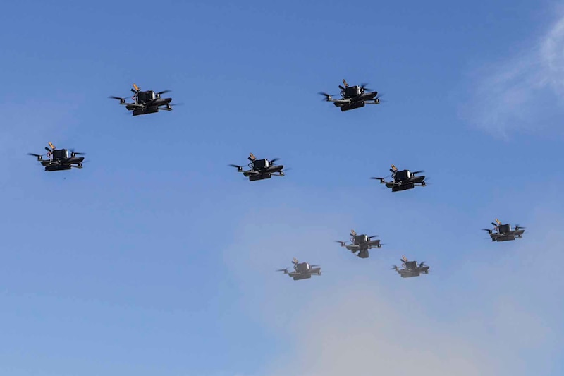 Nine drones fly as a group, with a blue sky and wispy clouds in the background.