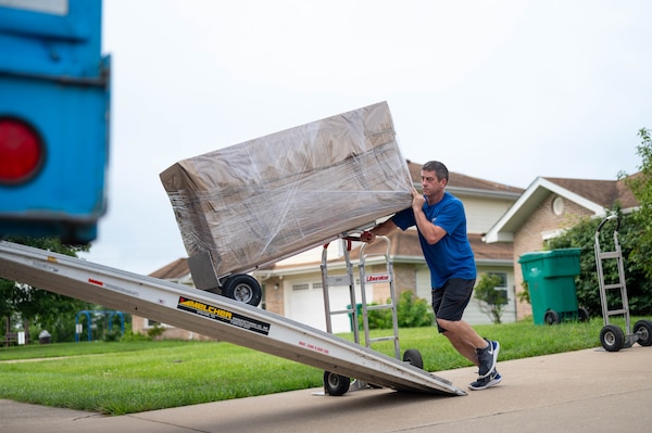 A person in casual attire pushes a hand truck with a package up a ramp that leads into a moving truck. There is a house in the background.