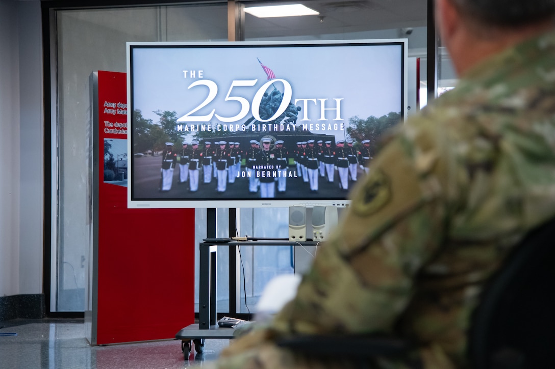 A person in a camouflage military uniform looks at a television screen that says 'the 250th Marine Corps birthday message' on it.