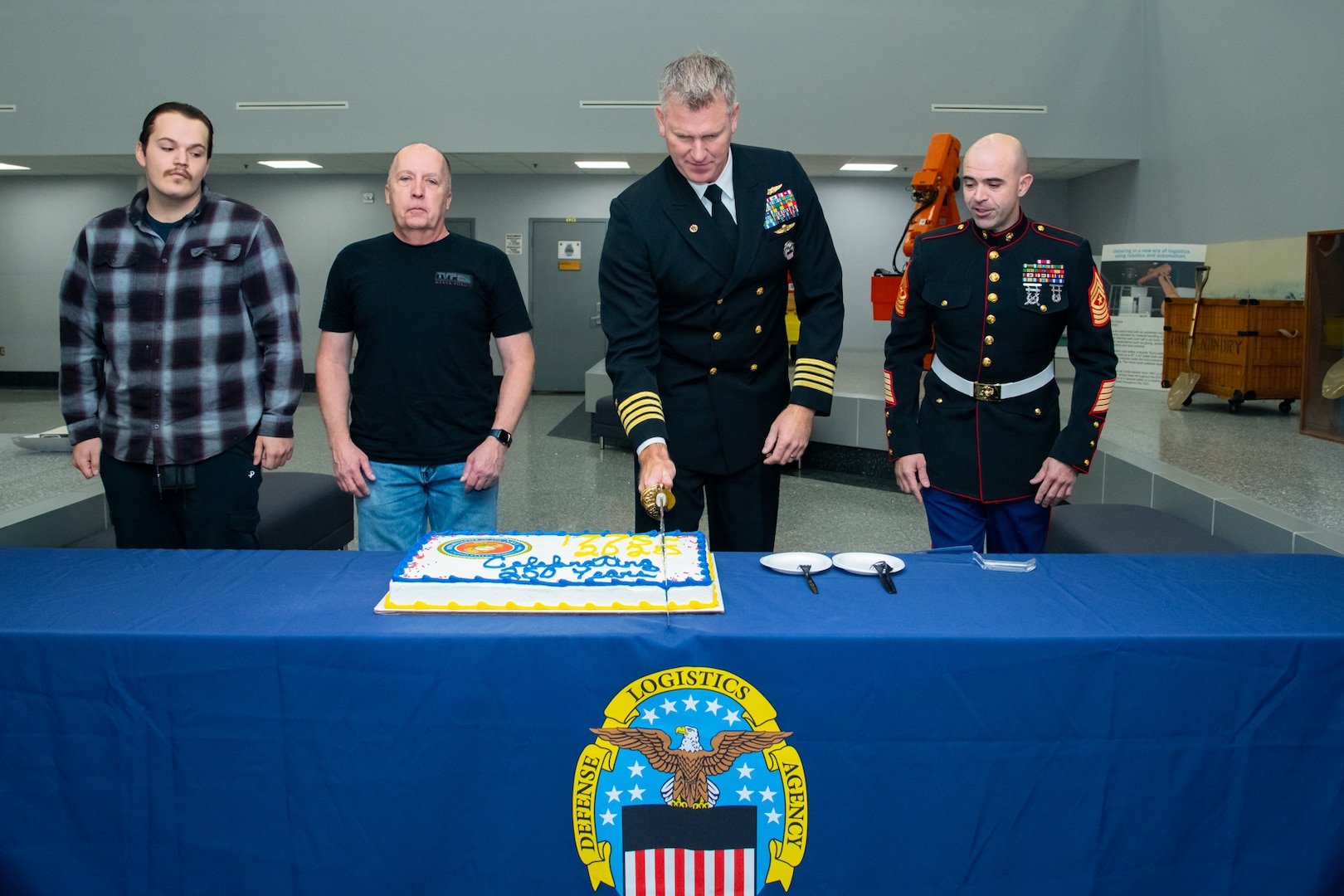 Four men stand behind a table with a sheet cake on it. Two of them are wearing military uniforms and one of the uniformed men is cutting the cake.