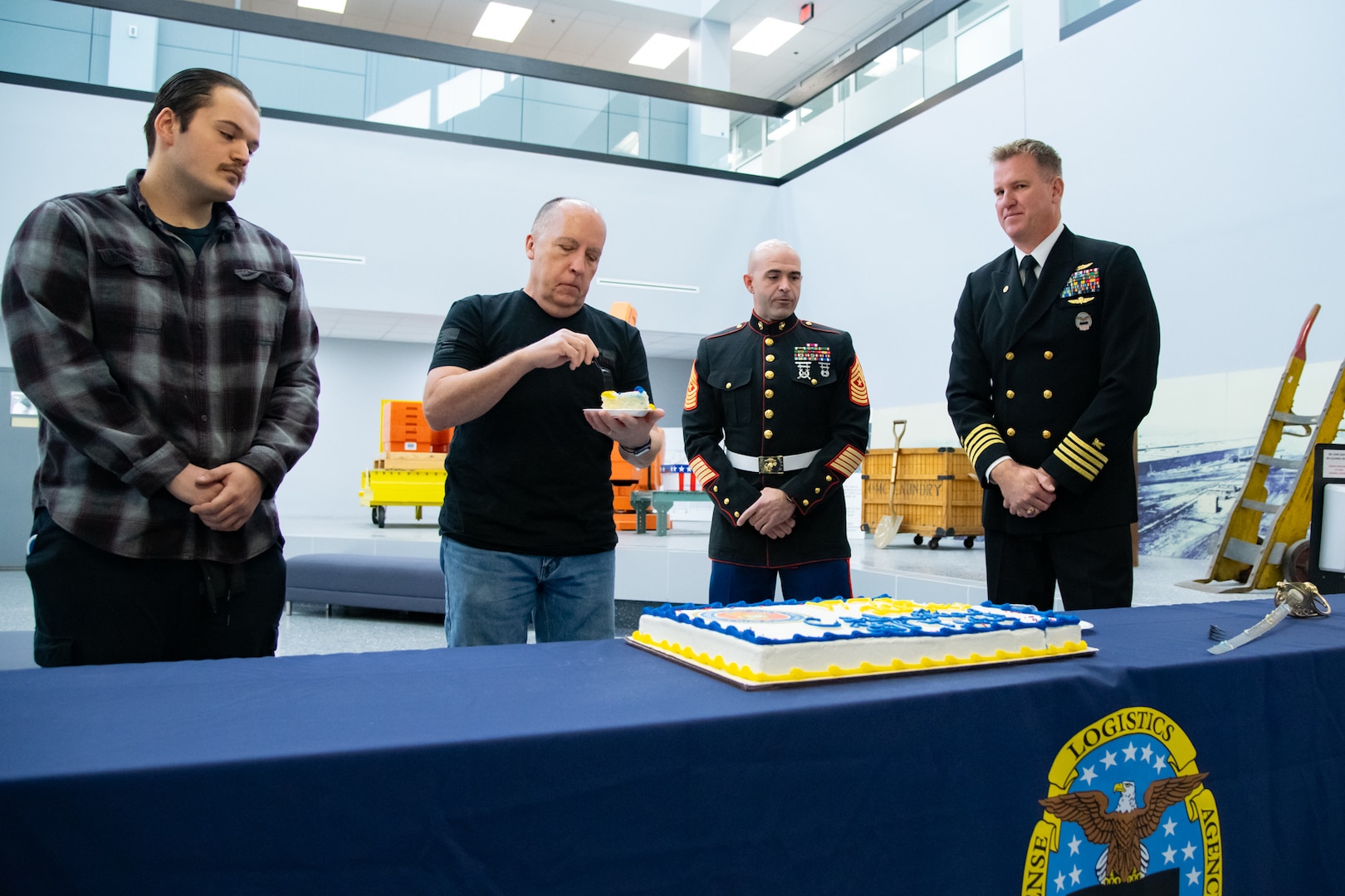 Four men stand behind a table with a sheet cake on it with one of them holding a piece of cake. Two of the men are wearing military uniforms.