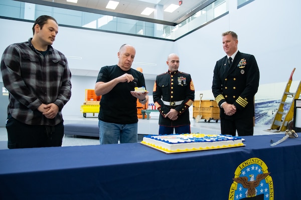 Four men stand behind a table with a sheet cake on it with one of them holding a piece of cake. Two of the men are wearing military uniforms.