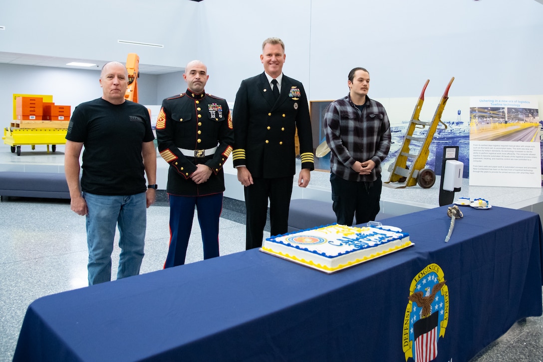 Four men stand behind a table with a sheet cake on it. Two of them are wearing military uniforms.