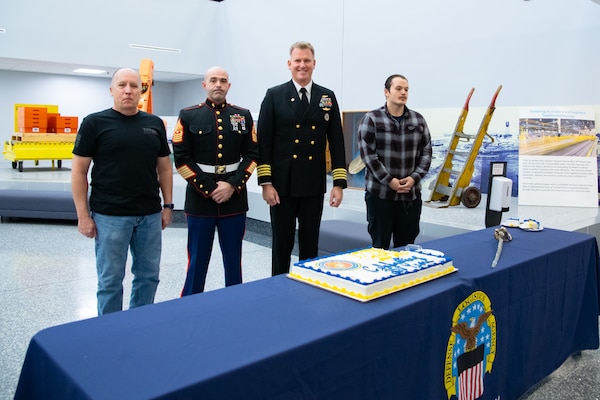 Four men stand behind a table with a sheet cake on it. Two of them are wearing military uniforms.