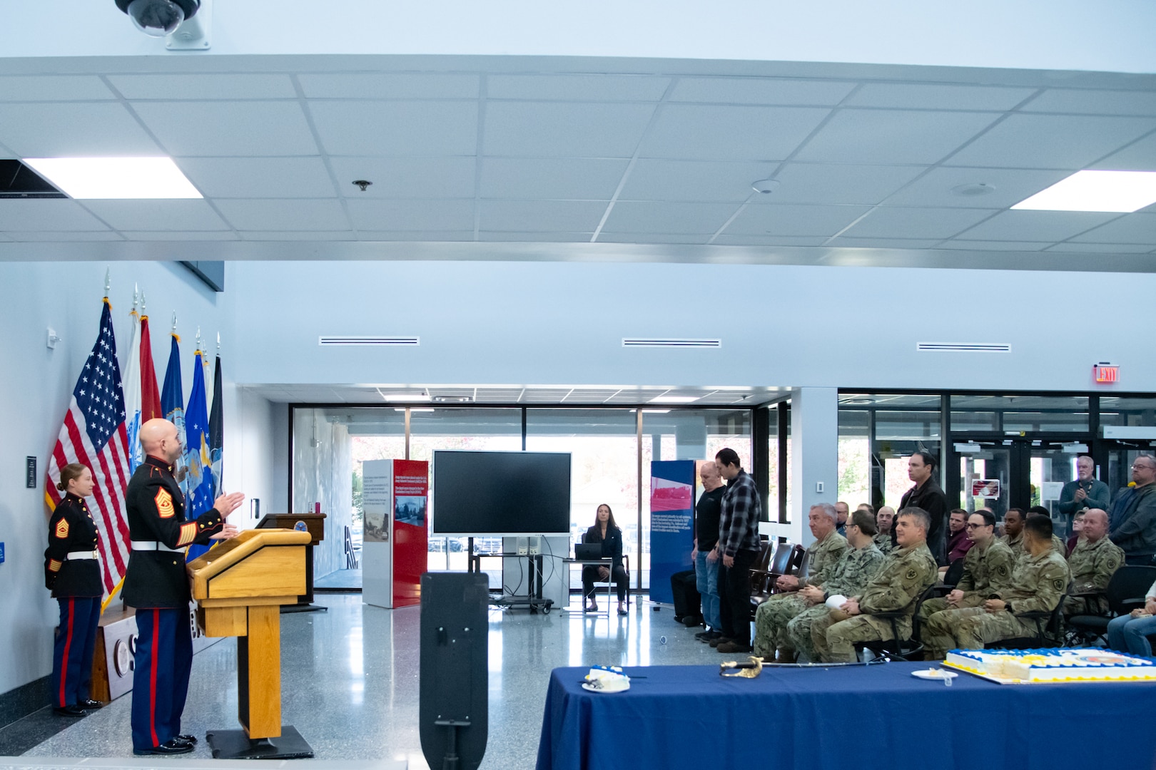 A man wearing a military uniform stands inside a building lobby behind a lectern speaking to a small audience.