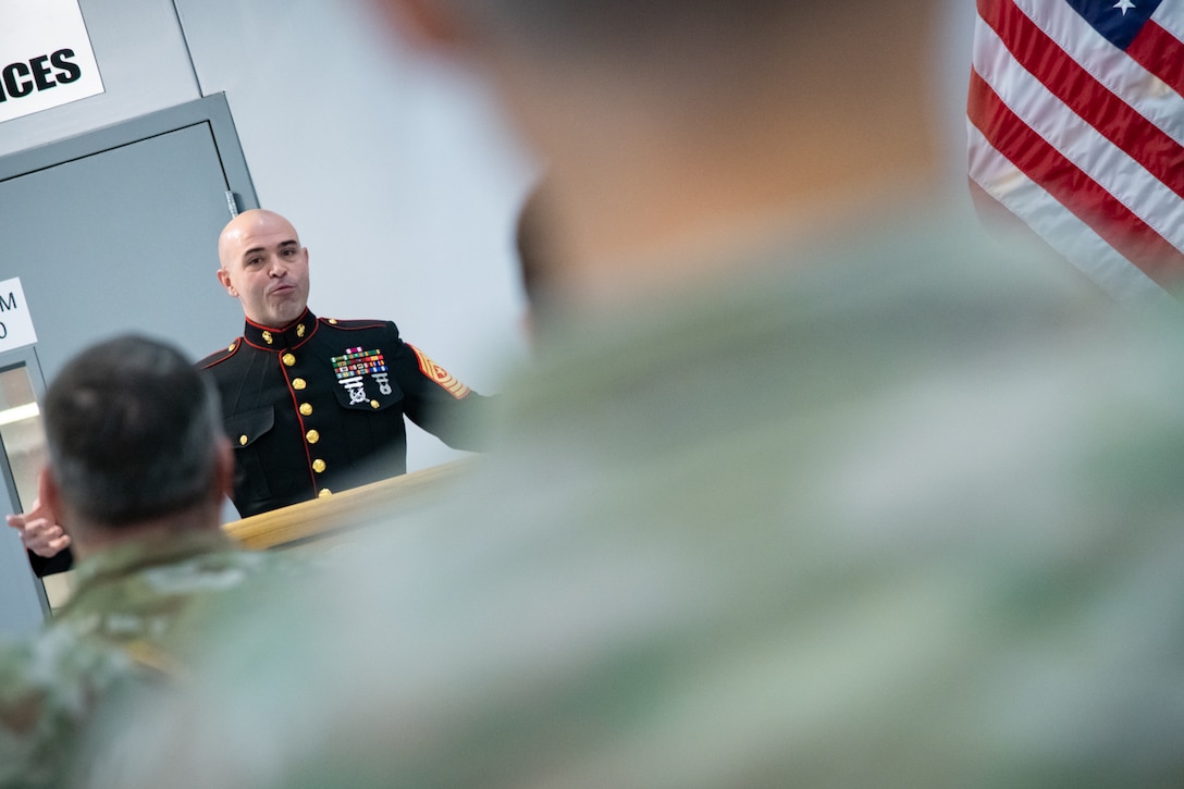 A man wearing a military uniform stands behind a lectern speaking to a group of people.