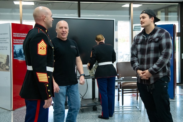 Three men stand inside of a building lobby speaking to each other, one is wearing a military uniform.