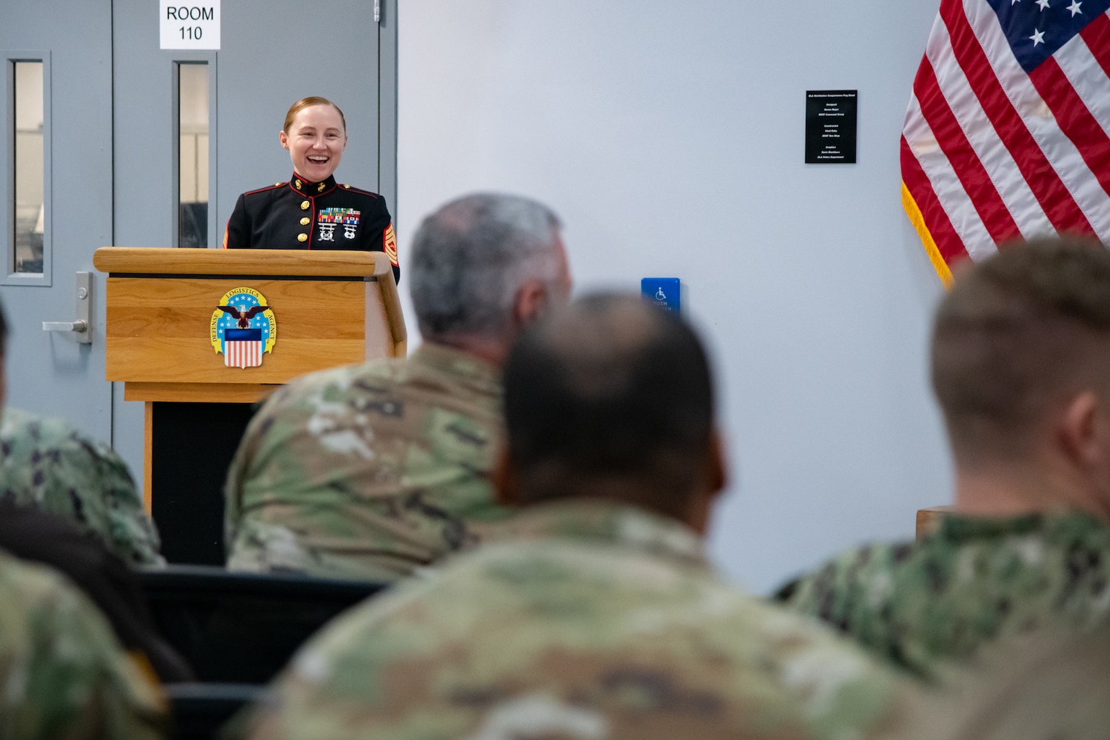 A woman wearing a military uniform stands inside behind a lectern speaking to a group of people sitting in the audience.