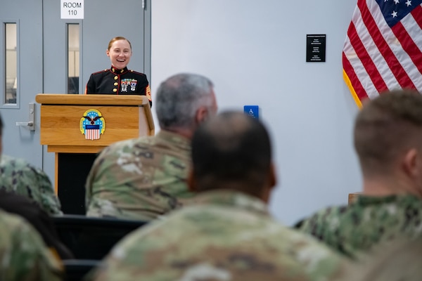 A woman wearing a military uniform stands inside behind a lectern speaking to a group of people sitting in the audience.