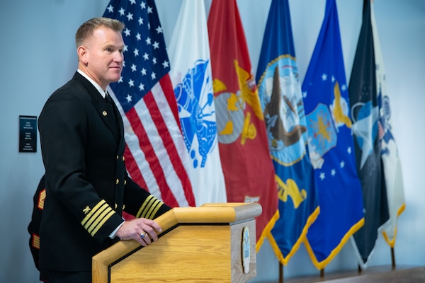 A man wearing a military uniform stands inside behind a lectern speaking.