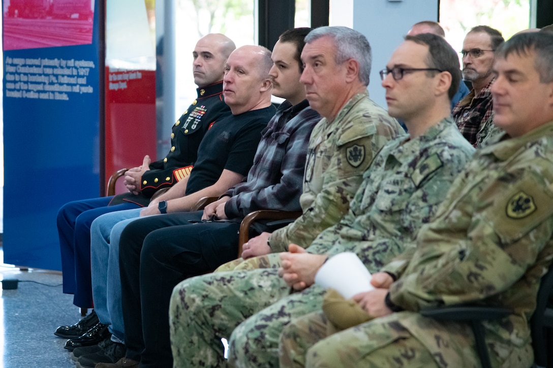 A group of people, most of them wearing camouflage military uniforms, sit inside a building lobby looking toward a person speaking.