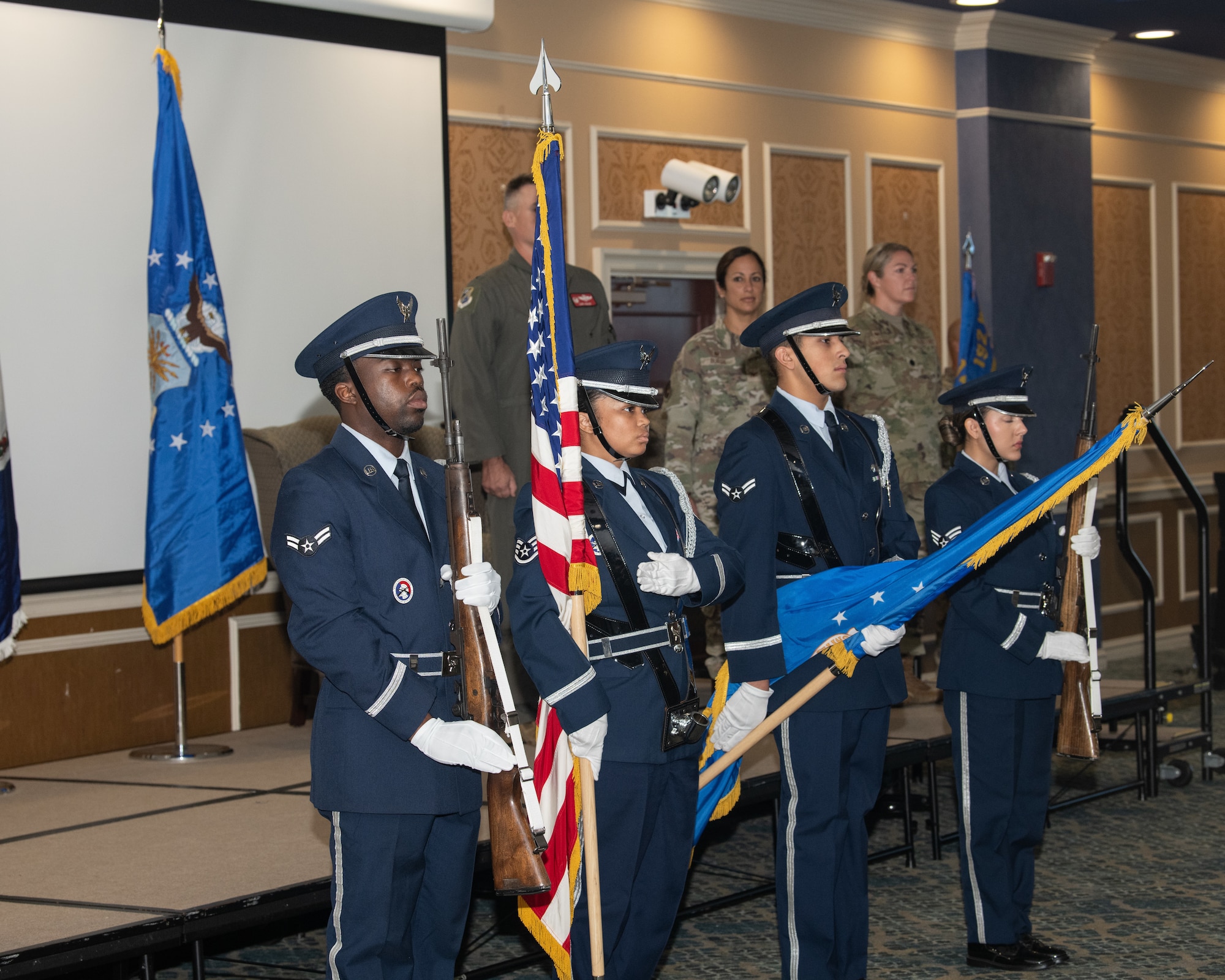A four-member U.S. Air Force honor guard stands in formation holding rifles and flags during a ceremony. Three USAF members stand at attention on the stage behind them.