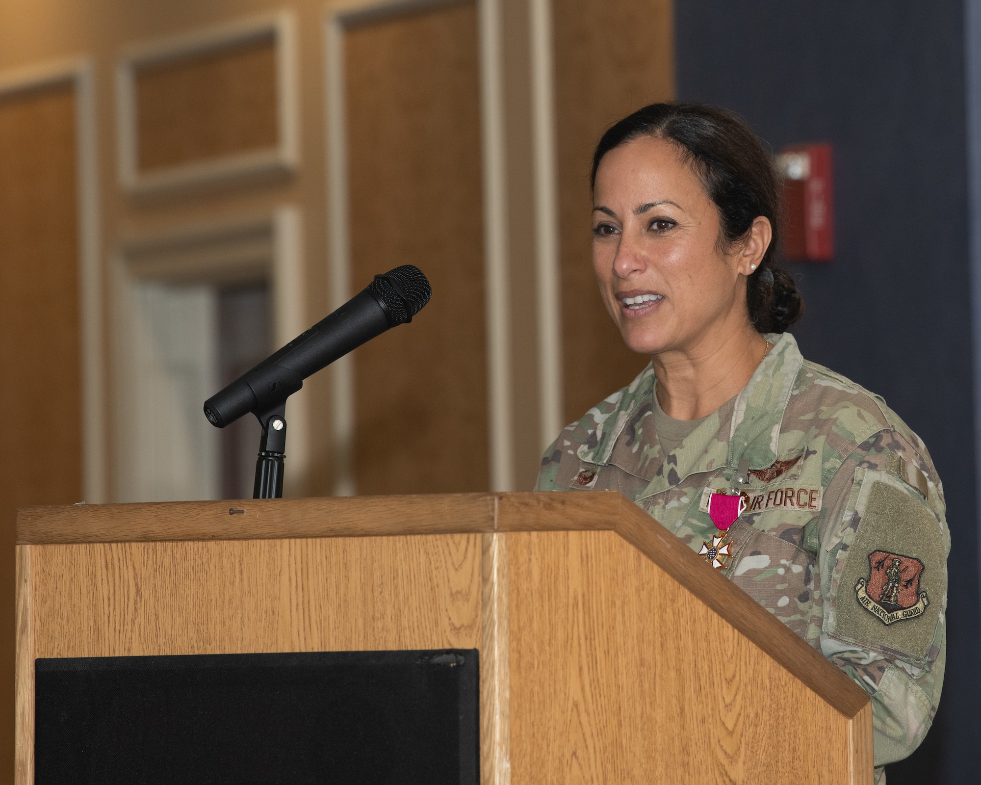Col. Manaal I. Burge stands at a wooden podium, speaking into a microphone. She is wearing a U.S. Air Force OCP uniform with a U.S. Meritorious Service medal pinned to her chest and is addressing an audience indoors.