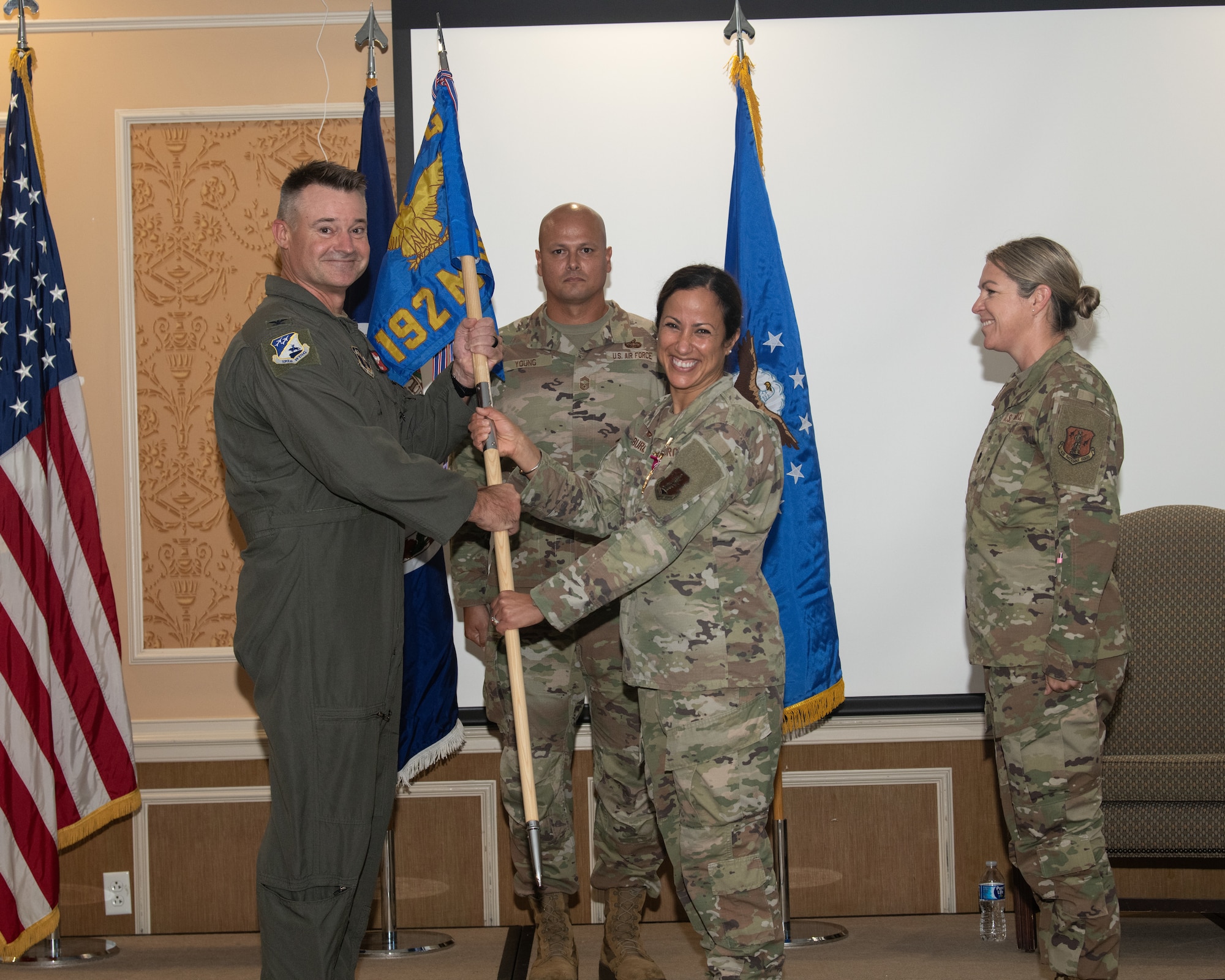 A U.S. Air Force change of command ceremony shows two uniformed service members smiling as they jointly hold a unit guidon. Another service member stands centered behind them, and a fourth stands to the right observing the moment. U.S. and unit flags are displayed in the background.