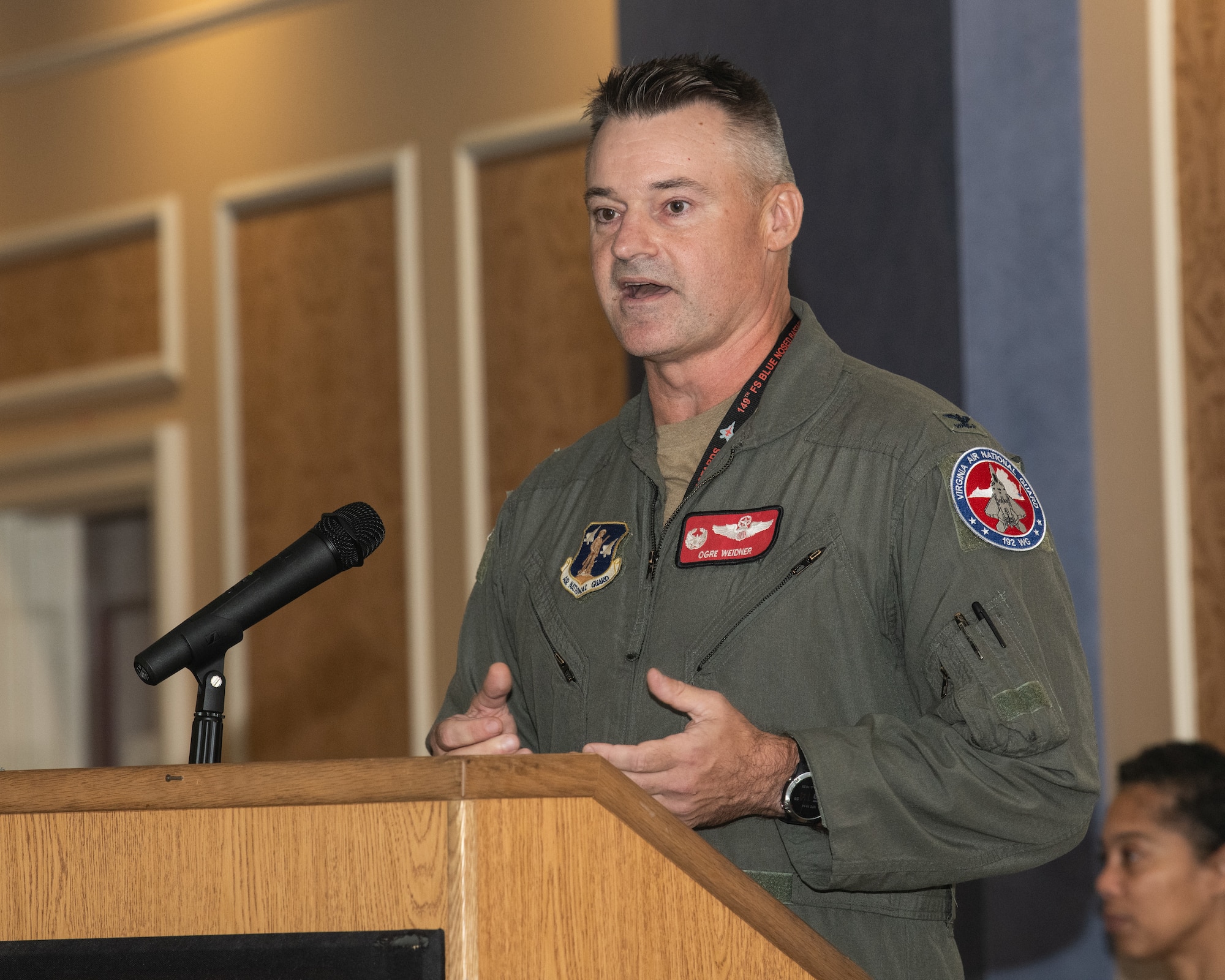 A U.S. Air National Guard member, Col. Andrew Weidner, speaks at a podium with a microphone, gesturing with his hands during a ceremony.