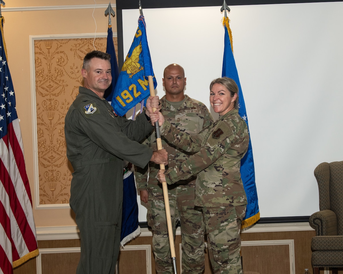 Two U.S. Air Force service members, Col. Andrew Weidner and Lt. Col. Jessica Gambone, hold a blue 192nd MSG unit guidon together during a ceremonial passing of the flag, while another service member stands at attention behind them.