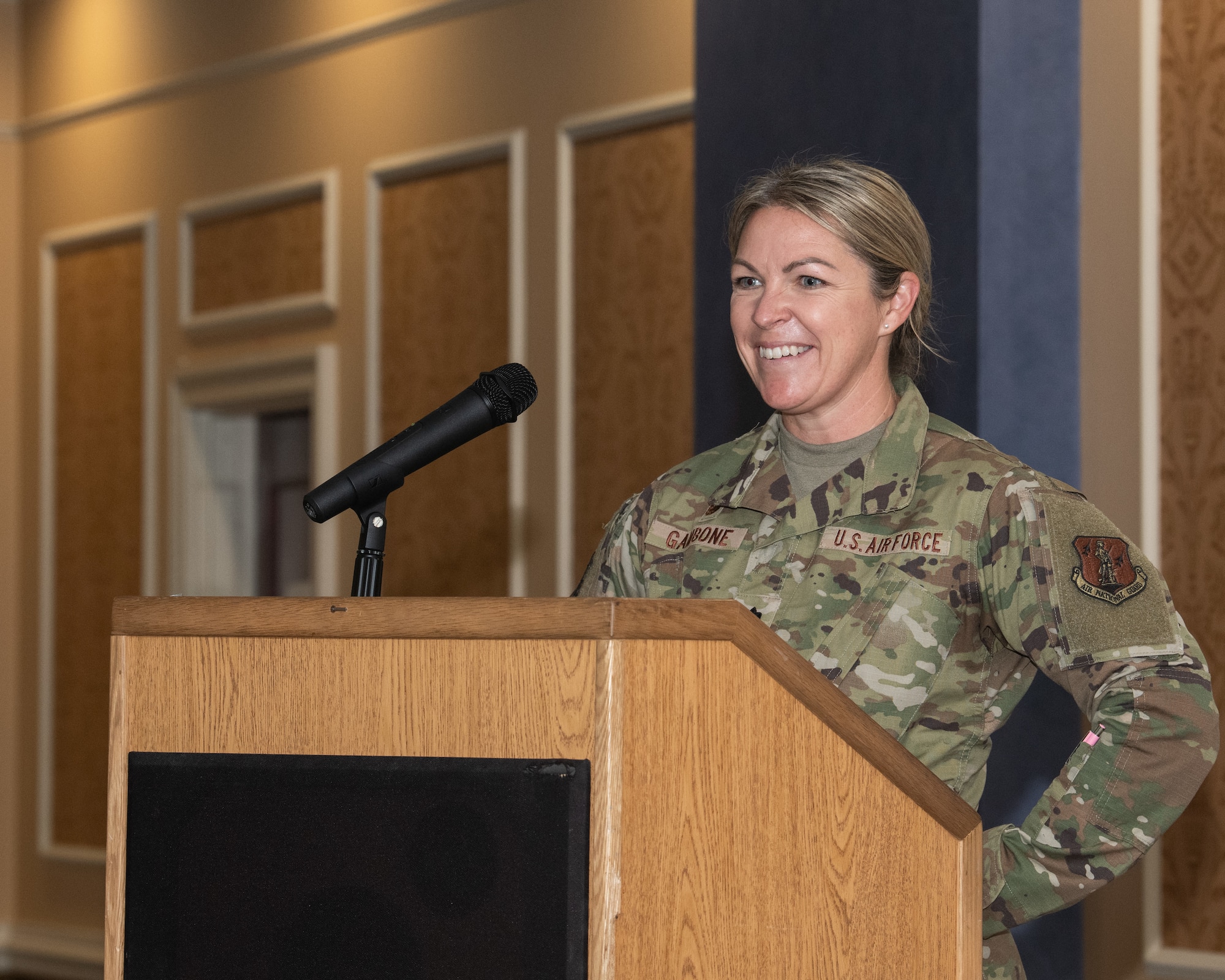 A U.S. Air National Guard member, Lt. Col. Jessica M. Gambone, smiles while speaking at a podium with a microphone during a ceremony.