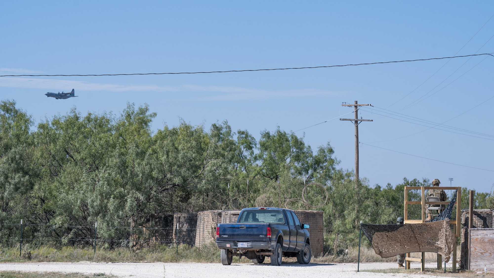 A U.S. Air Force C-130J Super Hercules assigned to the 317th Airlift Wing flies near Ray Rangel Air Base while Airmen monitor entry control points during exercise Coyote Shield at Dyess Air Force Base, Texas, Oct. 16, 2025. Coyote Shield enabled the 7th Civil Engineer Squadron and 7th Security Forces Squadron Airmen to enhance their expertise in base recovery, support reception, and command re-establishment, bolstering the 7th Bomb Wing's operational effectiveness in contested environments. (U.S. Air Force photo by Airman 1st Class Caleb Schellenberg)