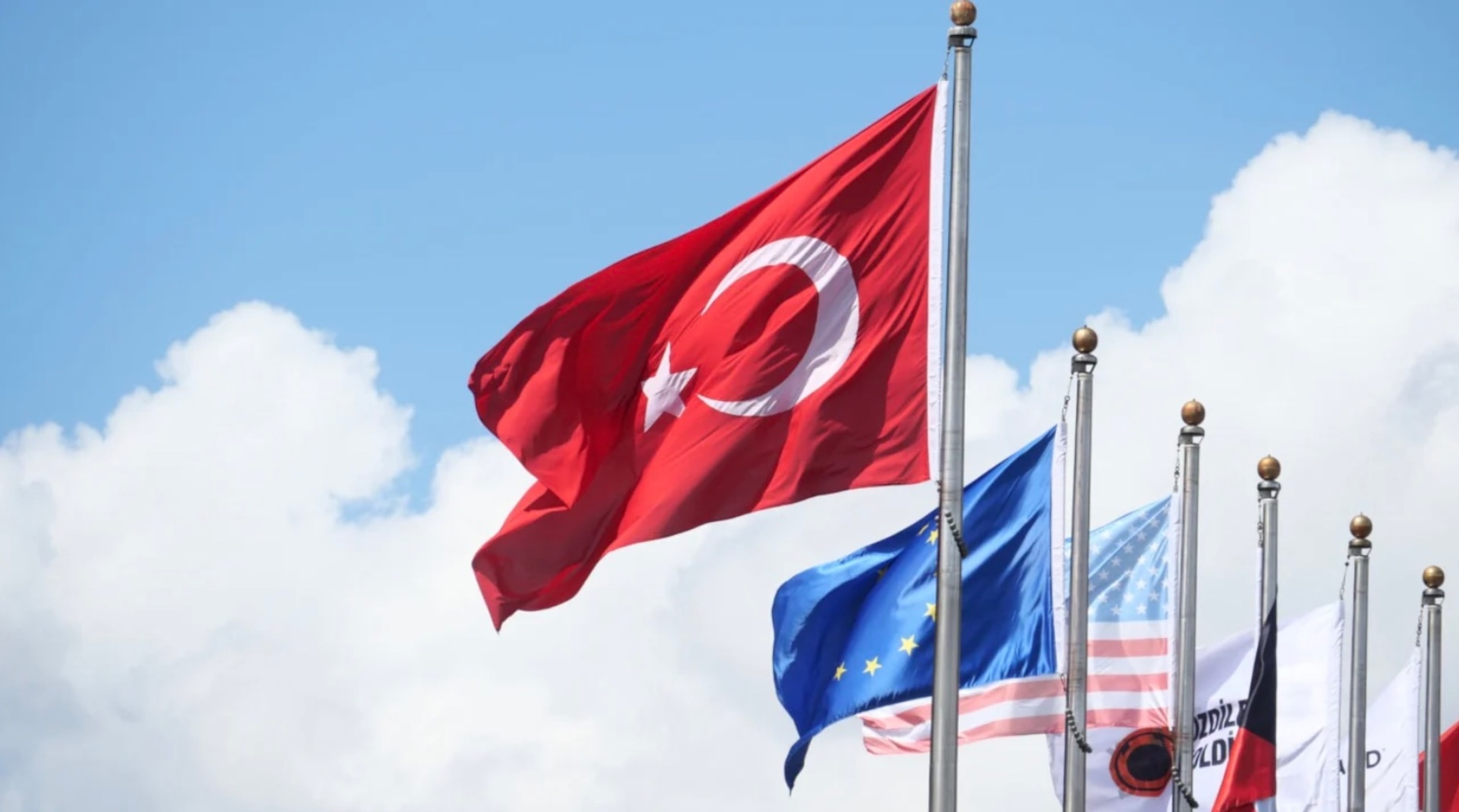 Multiple flags flying on tall flagpoles against a blue sky with clouds, with the Turkish flag prominently in the foreground and the European Union, United States, and other national and organizational flags visible in the background.