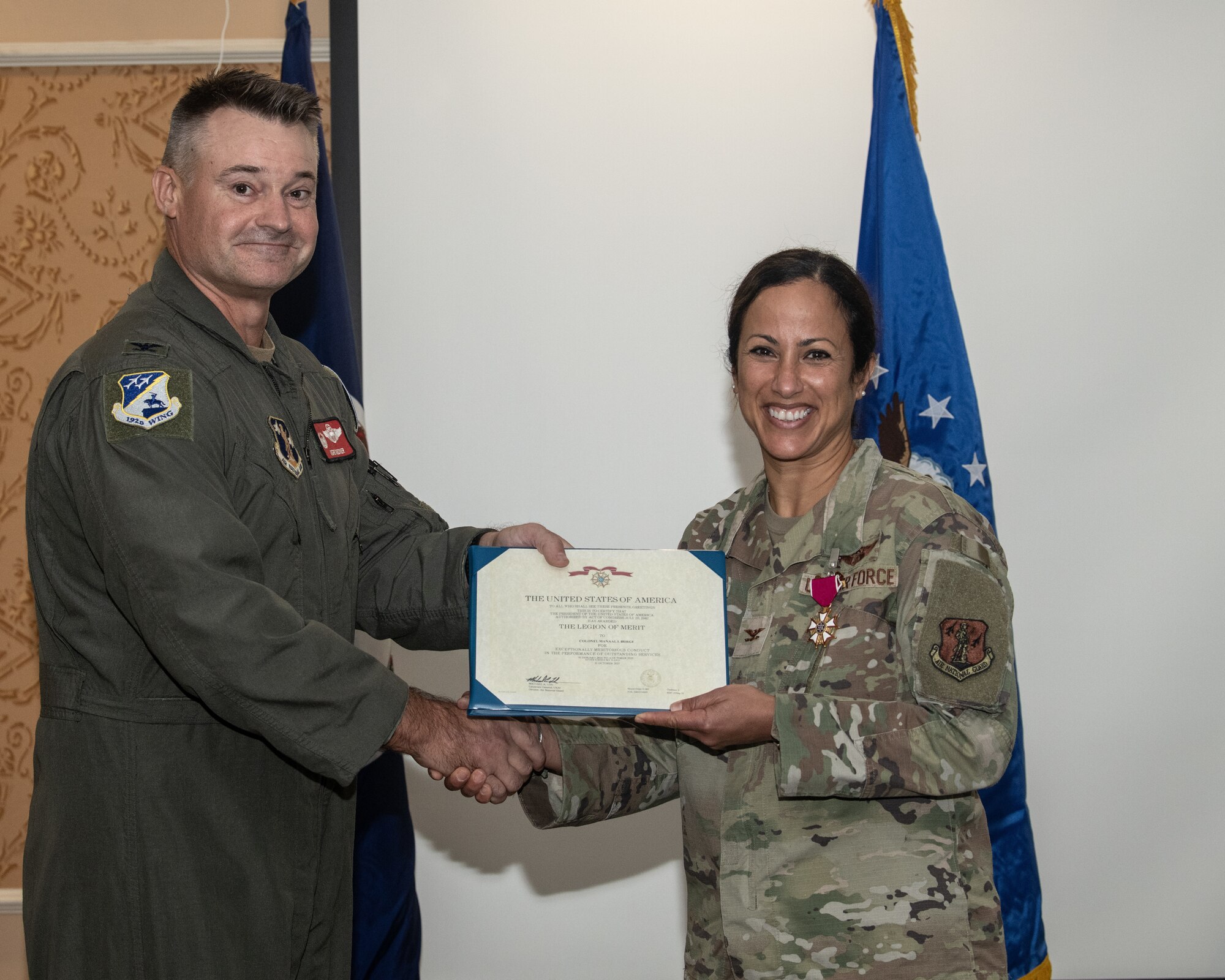 A U.S. Air National Guard member in a flight suit, Col. Andrew M. Weidner, presents a framed certificate for the U.S. Legion of Merit to another uniformed service member wearing USAF OCPs, Col. Manaal I. Burge, with the medal on her chest. The two shake hands and smile as they stand in front of a unit flag.