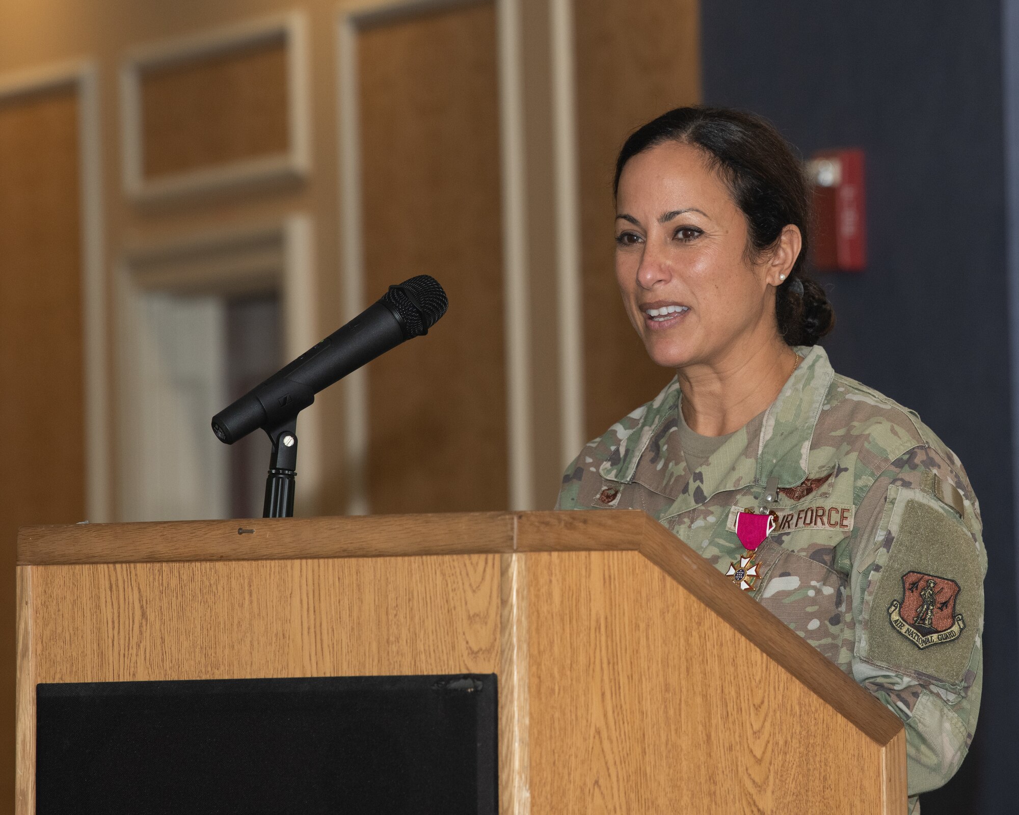 Col. Manaal I. Burge stands at a wooden podium, speaking into a microphone. She is wearing a U.S. Air Force OCP uniform with a U.S. Meritorious Service medal pinned to her chest and is addressing an audience indoors.