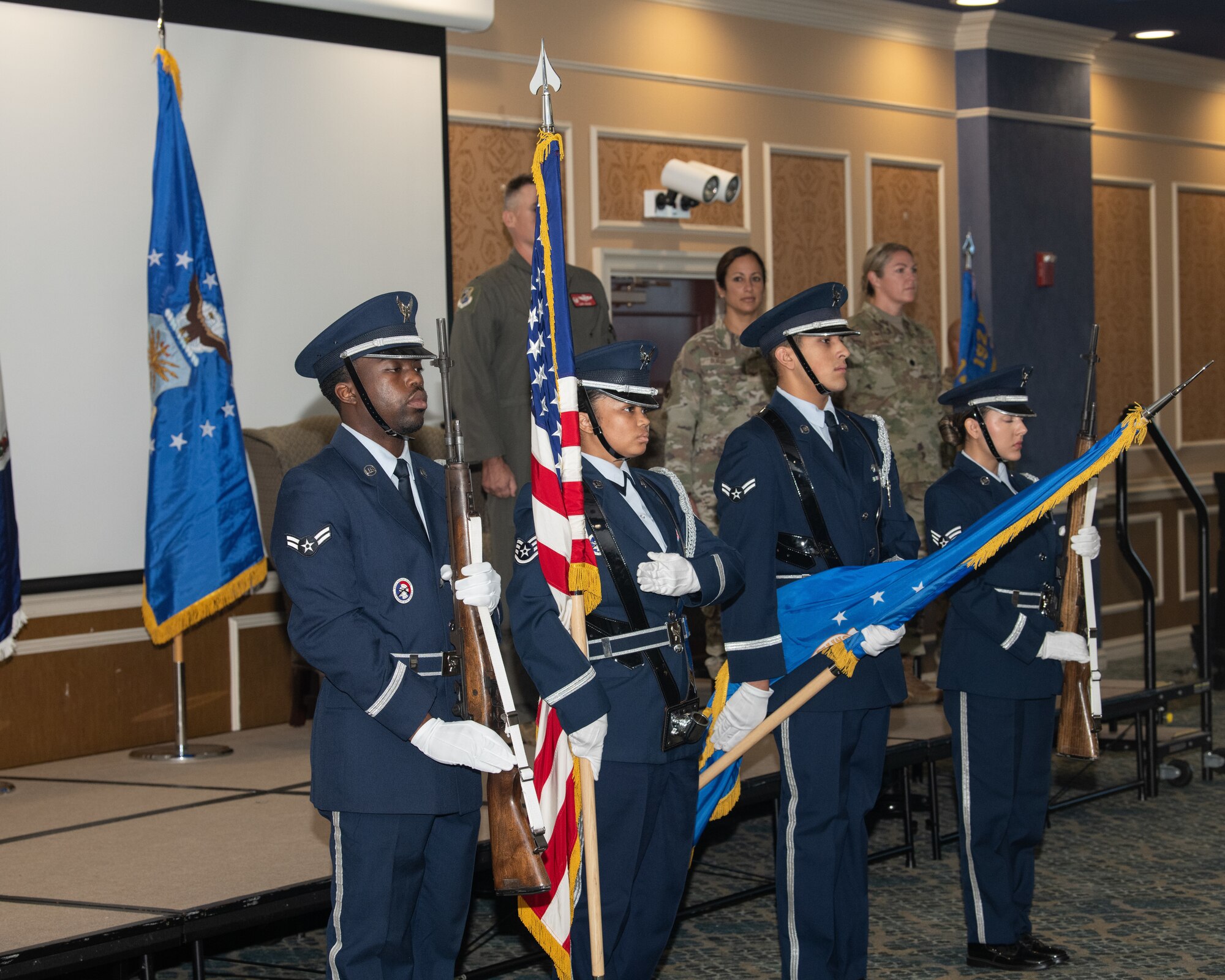 A four-member U.S. Air Force honor guard stands in formation holding rifles and flags during a ceremony. Three USAF members stand at attention on the stage behind them.