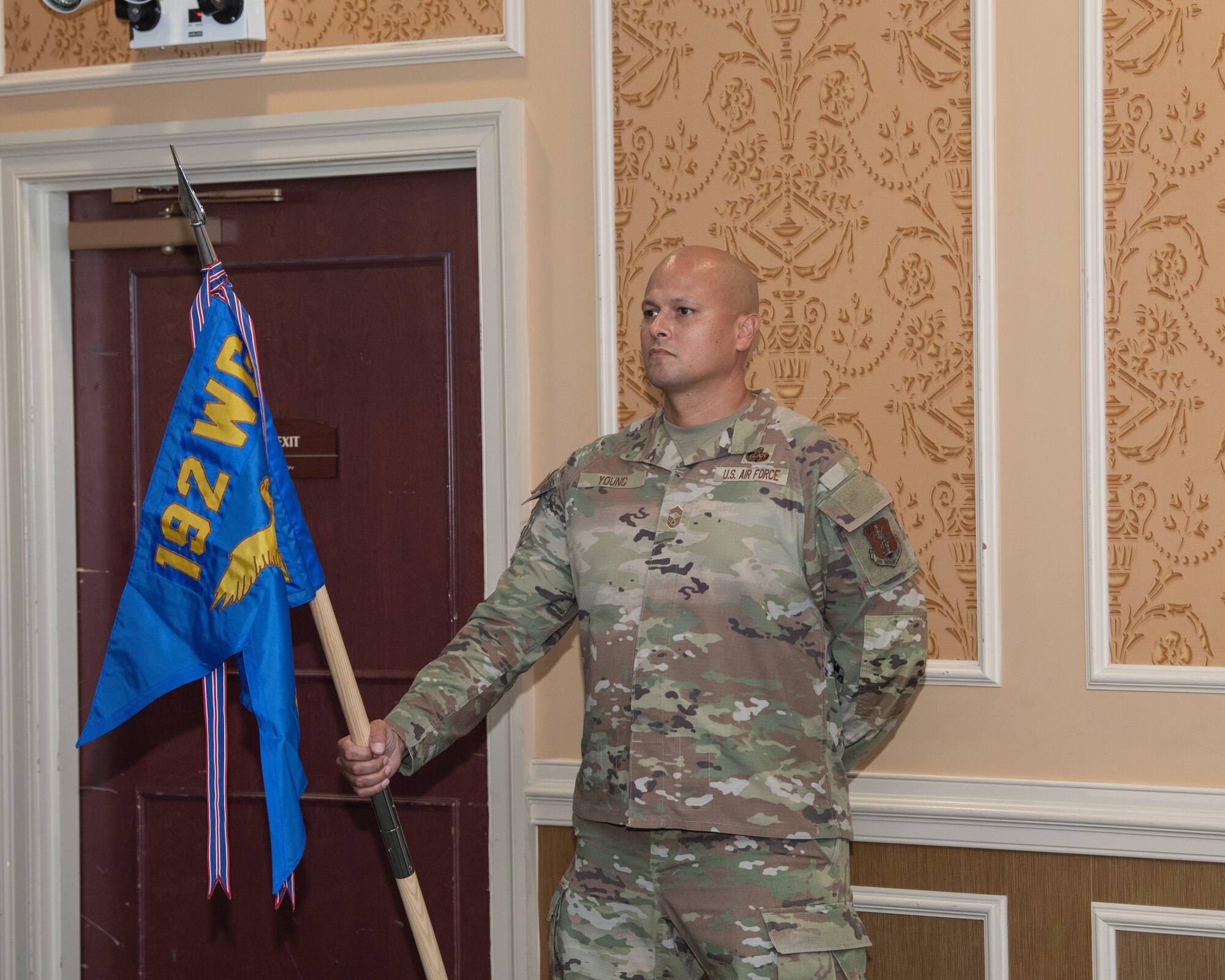 A U.S. Air National Guard member, Chief Master Sgt. Joseph Young stands at attention indoors, holding a blue 192nd MSG unit guidon flag.