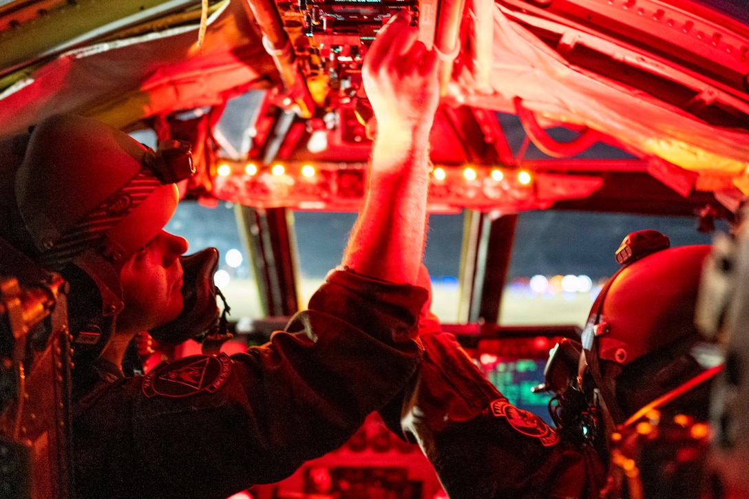 Two pilots sitting in the cockpit of an aircraft bathed in red light use their hands to adjust equipment, under dark conditions outside the plane.