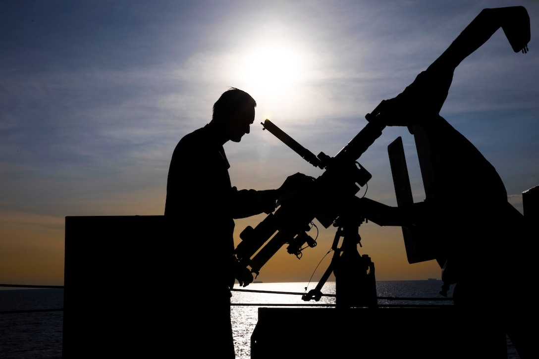 A sailor works on a large gun on the deck of a ship, silhouetted against an orange and blue sky and a body of water in the background.