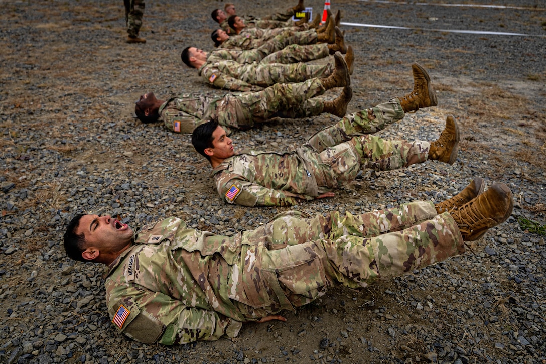 ROTC cadets in camouflage uniforms lie on their backs outdoors on the ground with their feet elevated.