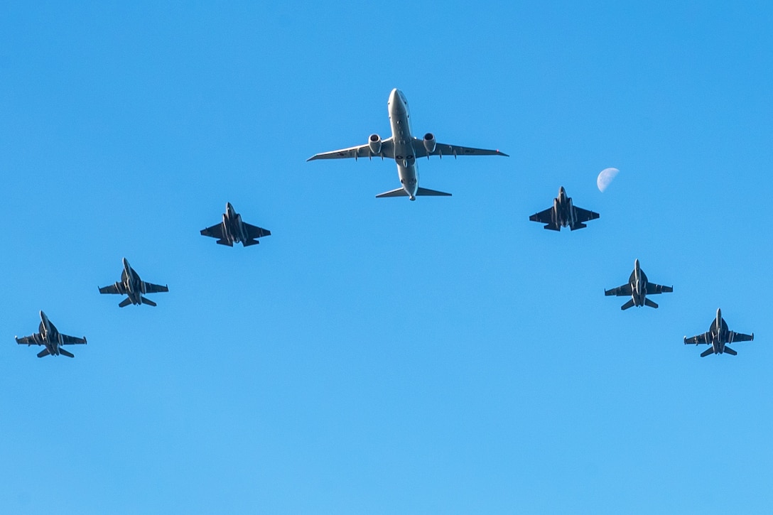 Six fighter jets and a larger aircraft fly in a V-formation on a cloudless day, with the moon in the background.