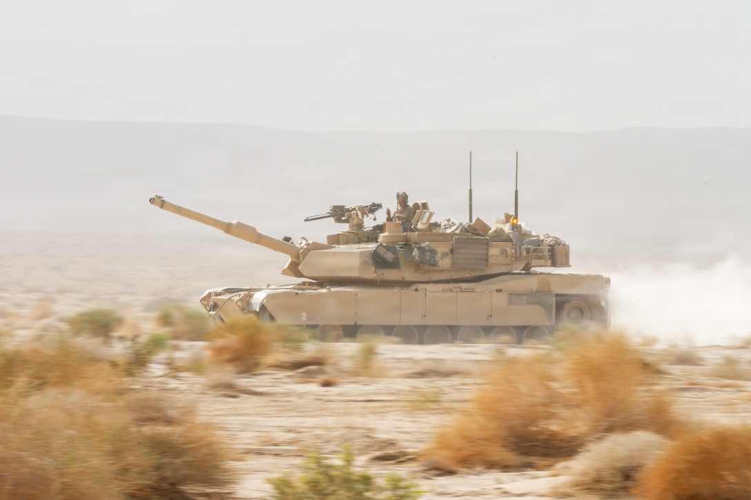 Soldiers operate a tank in sandy conditions during daytime, with hills in the background.
