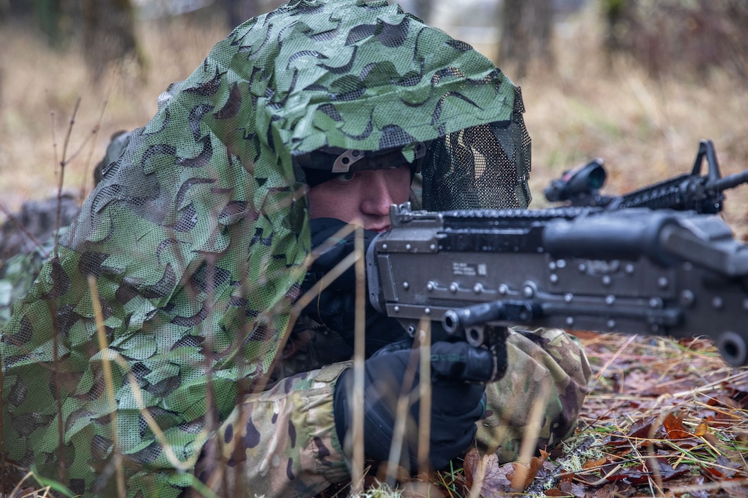 A soldier wearing camouflage and black gloves aims a rifle while lying on the ground in a forest setting during daytime.