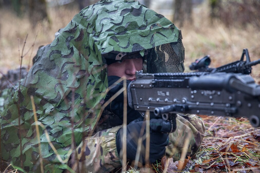 A soldier wearing camouflage and black gloves aims a rifle while lying on the ground in a forest setting during daytime.