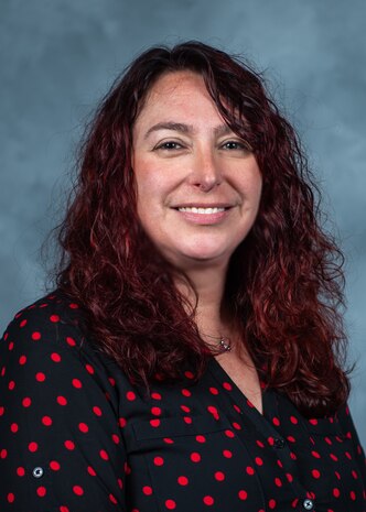 A portrait of a woman wearing a blue shirt with red polka dots, with wavy red hair past her shoulders, in front of a grey background.