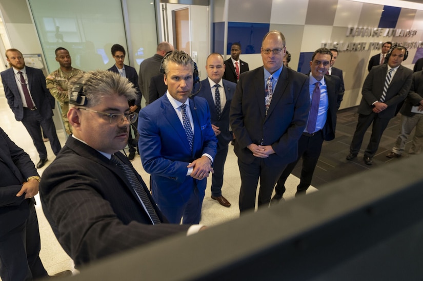More than a dozen men in business attire stand near each other and look at a television screen.