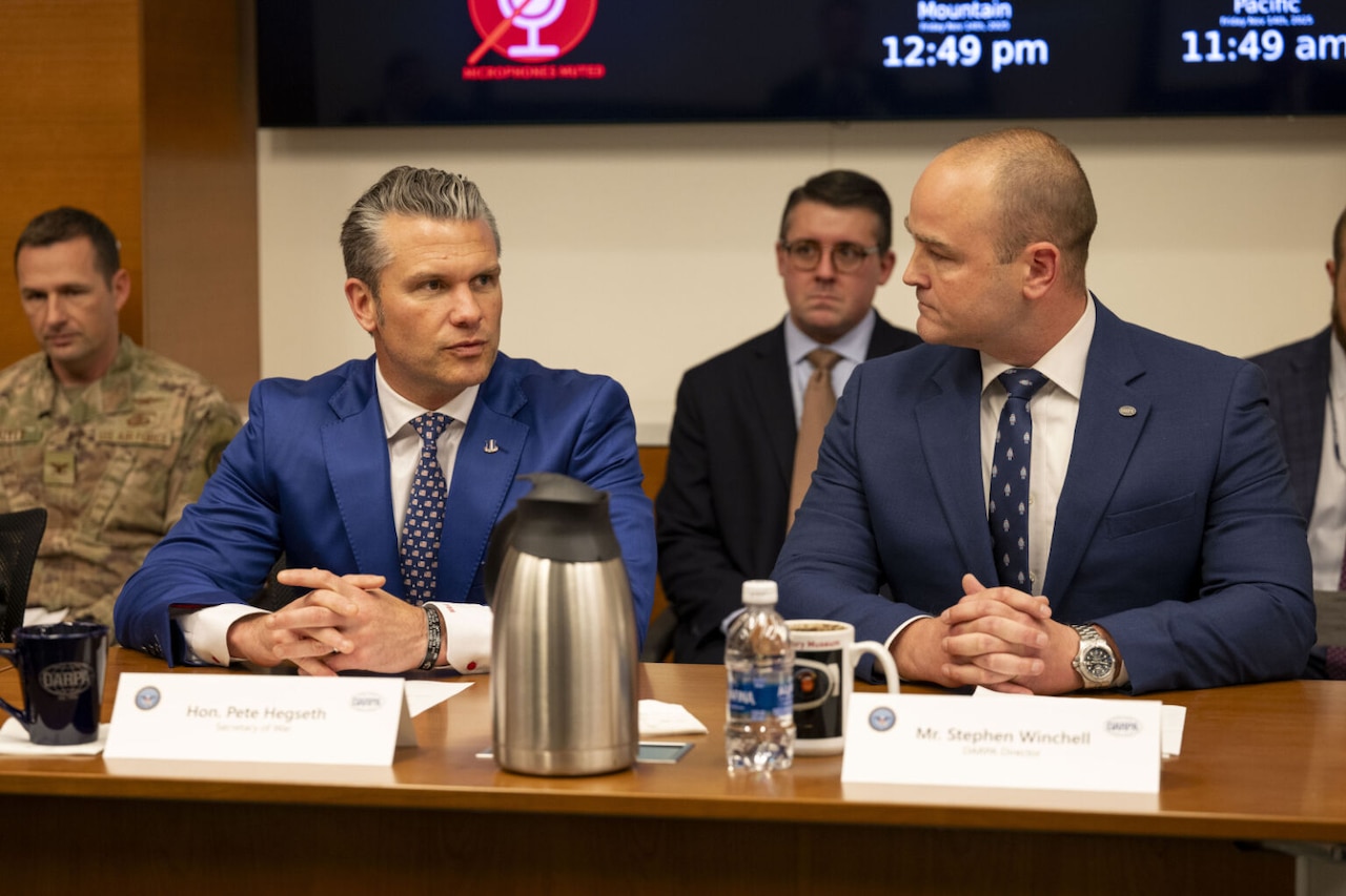 Two men in business attire are seated near each other at a table. Two other men in similar attire and one in a camouflage military uniform are seated behind them.