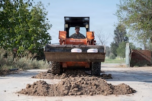 An Airman drives over two dirt hills in a skid steer loader with water buckets on the vehicle.
