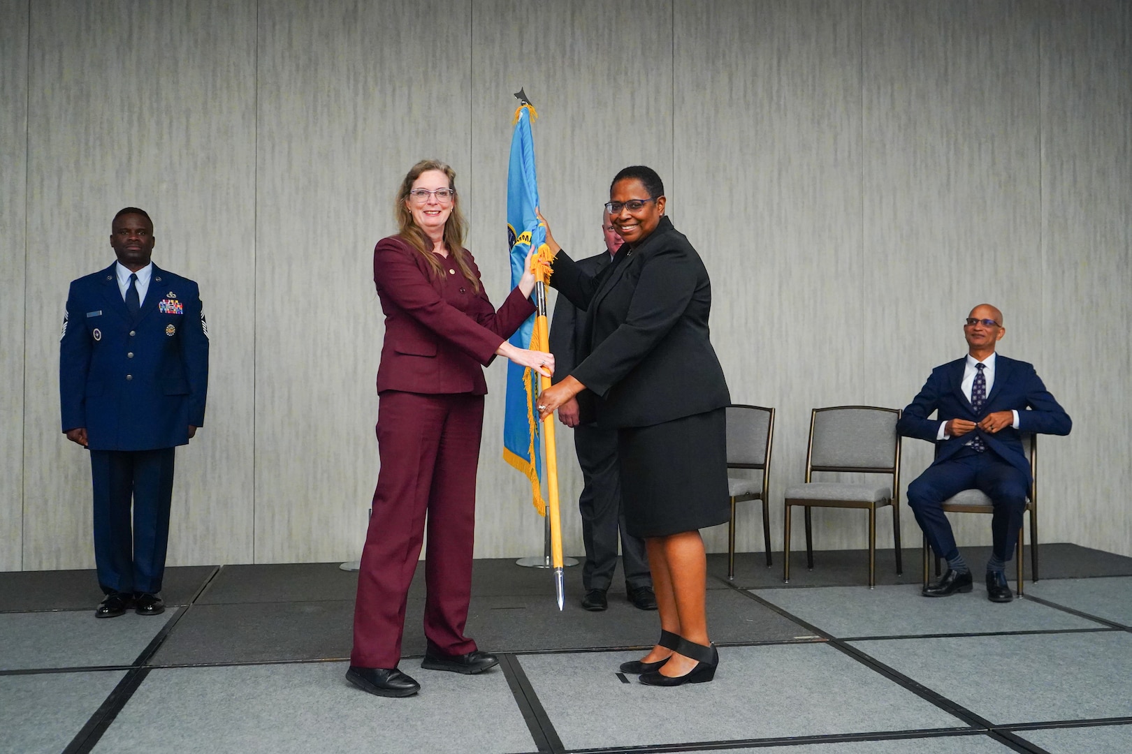 A woman wearing a burgundy pant suit hands a flag to a woman wearing a black suit