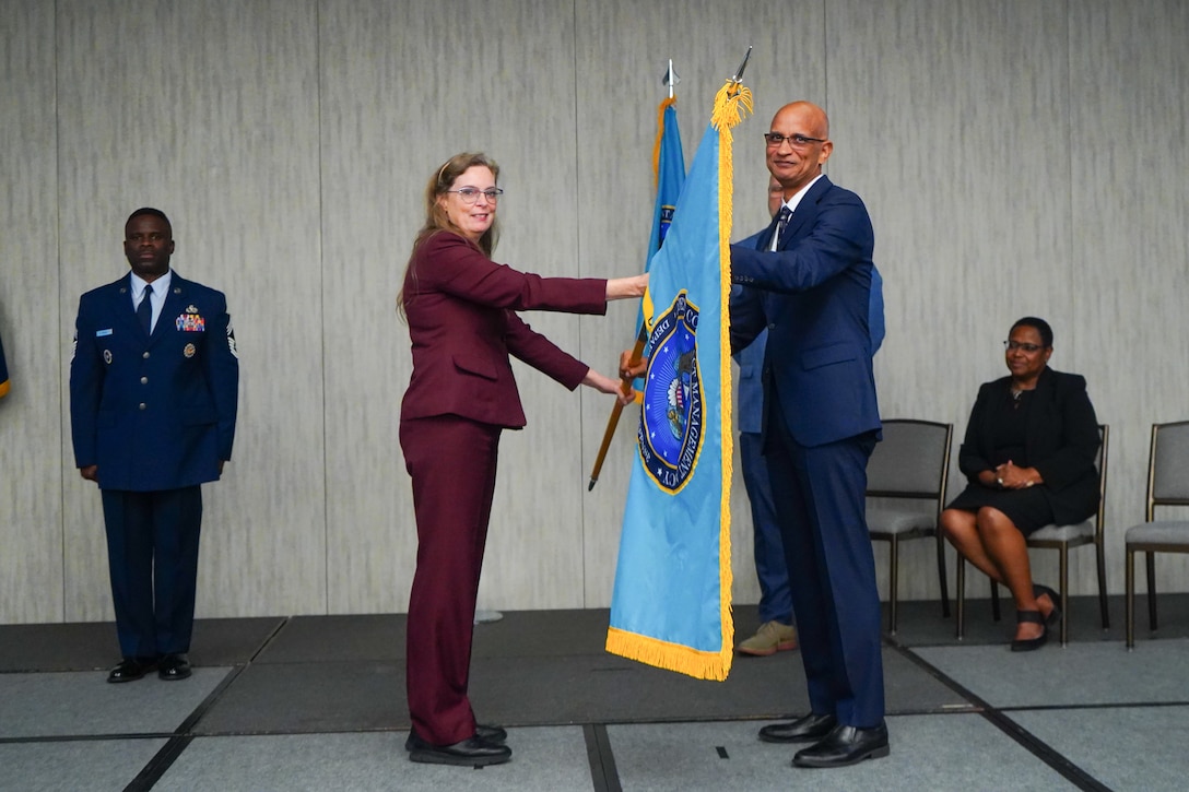 A woman wearing a burgundy pant suit hands a flag to a man wearing a blue suit