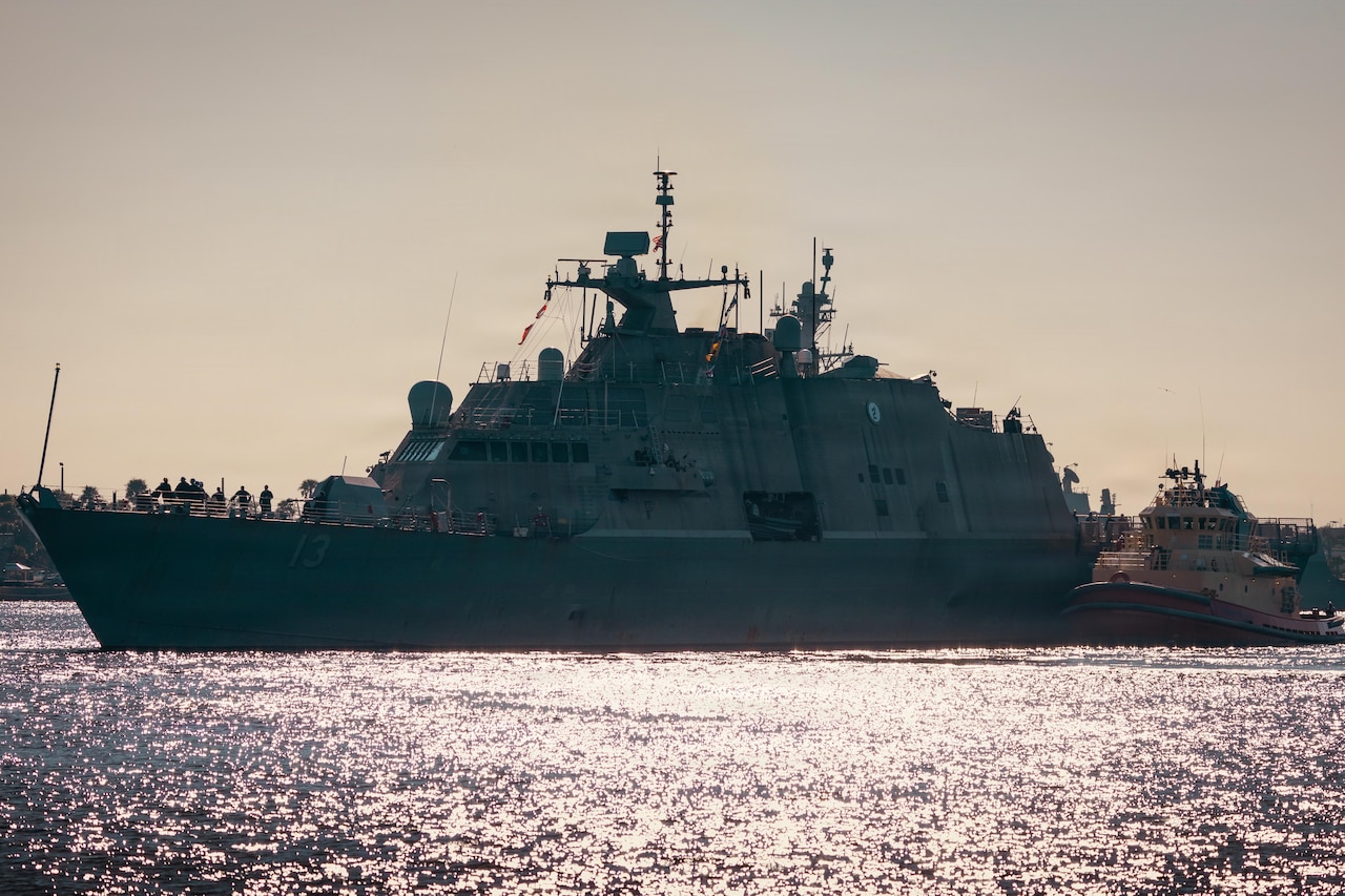A naval vessel navigates a body of water near land next to a smaller vessel.