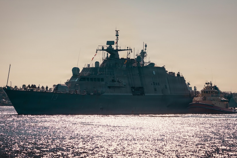 A naval vessel navigates a body of water near land next to a smaller vessel.