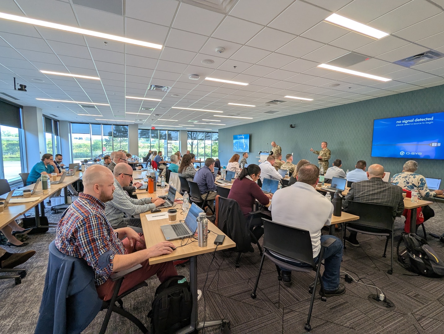 A group of men and women sit and listen as a man wearing a military uniform speaks to the group