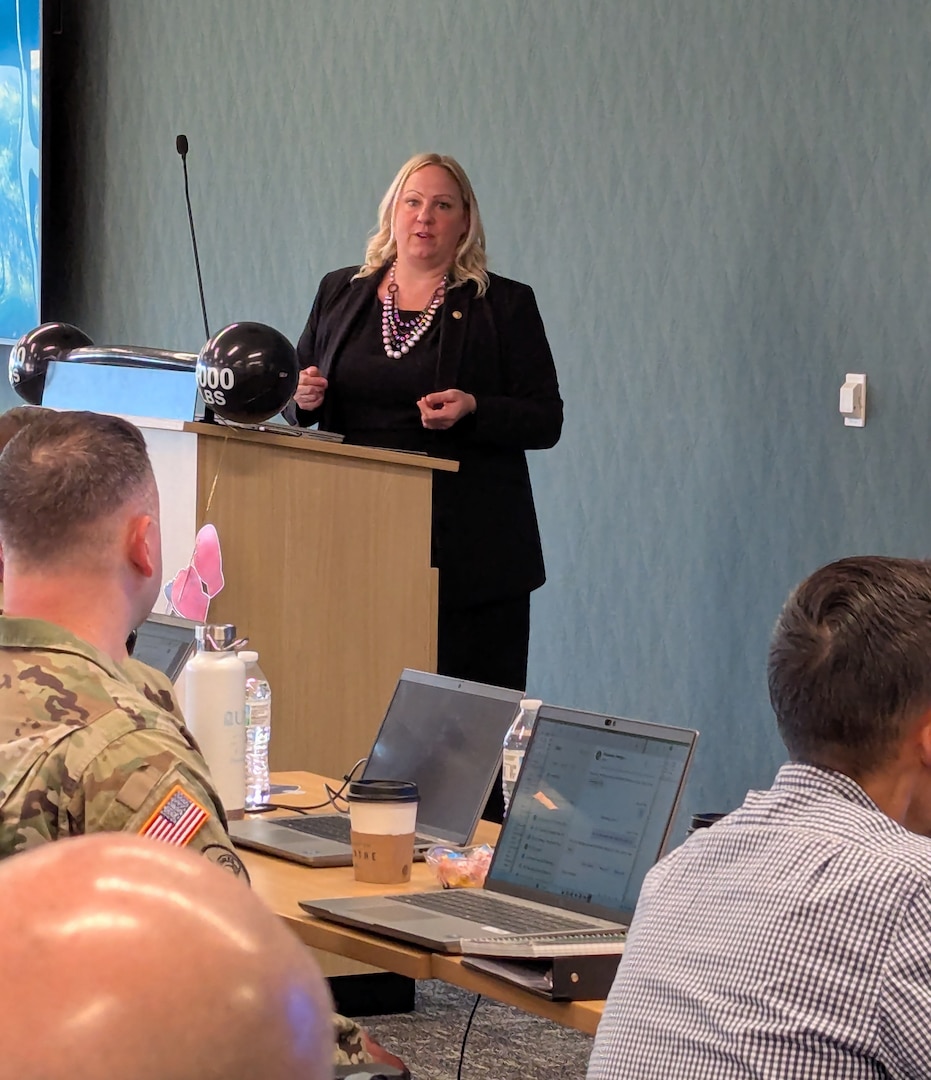 A woman wearing a black suit stands behind a podium and speaks to a group of men and women